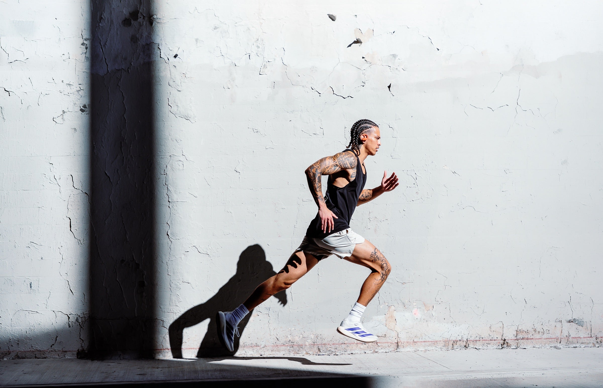 Runner in a black Haus tank and shorts sprinting past a white brick wall in hard sunlight